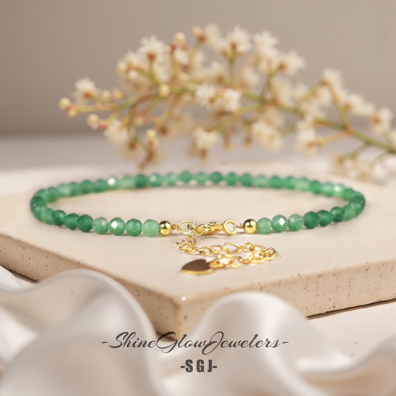 May include: A delicate green gemstone beaded bracelet with a gold heart charm and a gold clasp. The bracelet is on a white surface.