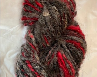 Op de afbeelding: Een bol garen in tinten rood, grijs en wit. Het garen is dik en pluizig, met een pluche textuur.
