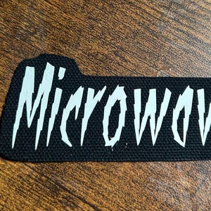 以下が含まれることがあります： 白いスタイリッシュな文字で「Microwave」と書かれた黒い布のパッチ