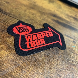 Puede incluir: Un parche Vans Warped Tour negro y rojo con una flecha blanca apuntando hacia la derecha. El parche está hecho de tela y tiene un borde cosido.