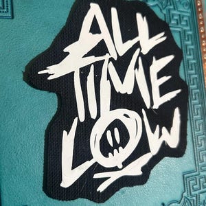Pode incluir: Patch de tecido preto com letras brancas que dizem "All Time Low" com um gráfico de smiley estilizado.