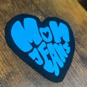 Könnte beinhalten: Ein schwarzer herzförmiger Patch mit den Worten "MOM JEANS" in blauer Schrift. Ein kleines Herz befindet sich im "O" von MOM.