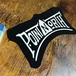 Può includere: Toppa ricamata in bianco e nero con il testo "POINT NORTH" in un font stilizzato.