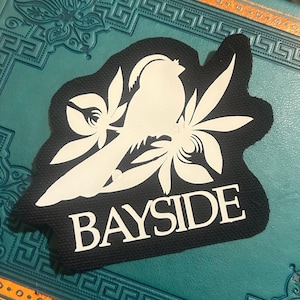May include: Black patch with a white bird and floral design. The word "BAYSIDE" is printed in white block letters below the bird. The patch is rectangular with rounded corners.
