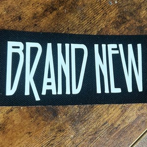 Pode incluir: Um patch retangular preto com as palavras "BRAND NEW" em letras maiúsculas brancas. O patch está sobre uma superfície de madeira com grão visível.