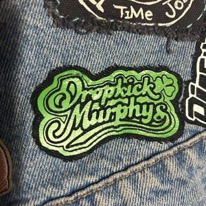 Peut inclure: Veste en jean ornée de plusieurs écussons, dont un vert et noir avec l'inscription stylisée "Dropkick Murphys". On peut également voir un écusson avec l'inscription "Full Time Job".