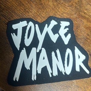 Puede incluir: Parche de tela negro con letras blancas que dicen "JOYCE MANOR" en una fuente estilizada y manuscrita.