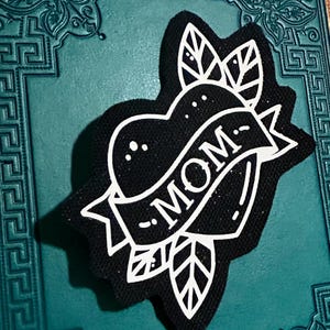 Könnte beinhalten: Schwarz-weißer herzförmiger Patch mit dem Wort "MOM" auf einem Banner. Das Herz ist von zwei Blättern umgeben.