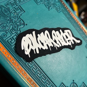 Op de afbeelding: Zwarte patch met witte graffiti-lettering die "PSYCHO KILLER" luidt. De patch bevindt zich op een turquoise boek met gouden accenten.