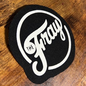 May include: A black and white circular patch with the text "THE FRAY" in a white cursive font.