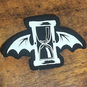 Puede incluir: Parche de tela negro con un reloj de arena blanco con alas. El reloj de arena está en un marco de madera y las alas se asemejan a las de un murciélago.
