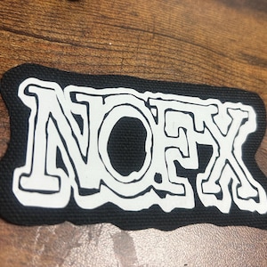 Pode incluir: Um remendo de tecido preto com a palavra "NOFX" escrita em branco, contornada de preto.