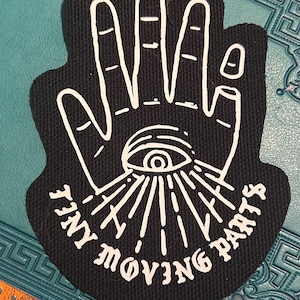 Könnte beinhalten: Schwarzer Stoff-Patch mit einem weißen Umriss einer Hand mit einem Auge in der Handfläche. Der Text "Tiny Moving Parts" ist um das Auge herum geschrieben.