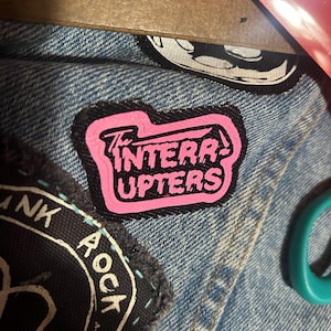 May include: A black and pink patch with the text "The Interrupters" on a denim jacket.