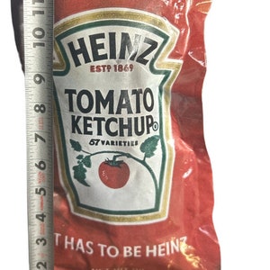 May include: A red Heinz Tomato Ketchup packet with the text "TOMATO KETCHUP" and "57 VARIETIES". The packet is approximately 6 inches tall. The text "Tear Here" is visible. The Heinz logo is at the top.