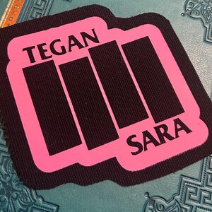 Könnte beinhalten: Rosa Stoff-Patch mit schwarzem Text, der "TEGAN" und "SARA" lautet, mit einer schwarzen Silhouette von vier vertikalen Balken in der Mitte.