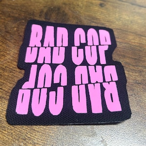 Peut inclure: Patch en tissu rose avec les mots "Bad God" écrits dans une police audacieuse et en blocs.