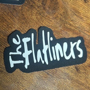 Puede incluir: Parche de tela negro con letras blancas que dicen "The Flatliners".