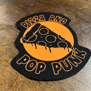 Könnte beinhalten: Schwarzer und orangefarbener gestickter Aufnäher mit dem Text "Pizza and Pop Punks" und einem Pizzastück mit tropfendem Käse.