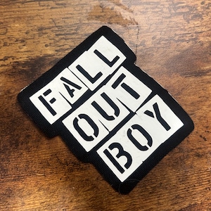 Może przedstawiać: Czarna naszywka z białym napisem "FALL OUT BOY" w stylu szablonowym. Naszywka ma czarną obwódkę i jest umieszczona na drewnianej powierzchni.