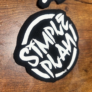 Op de afbeelding: Zwarte en witte geborduurde patch met de woorden "Simple Plan" in een gestileerd lettertype.