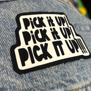 May include: A black and white patch with the text "Pick it up! Pick it up! Pick it up!!!" on a blue denim background.