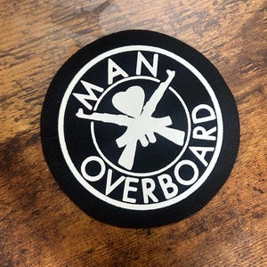 Op de afbeelding: Een zwarte ronde patch met witte tekst die "MAN OVERBOARD" luidt en een witte afbeelding van twee gekruiste geweren met een hart in het midden.