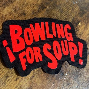 May include: A black patch featuring the text "Bowling for Soup!" in bold, red lettering. The patch has an unusual, curved outline. It is designed to be attached to fabric items, such as jackets or bags.