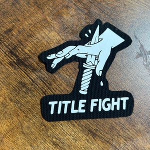 May include: Black and white patch with a hand holding a knife and the text "TITLE FIGHT" underneath.