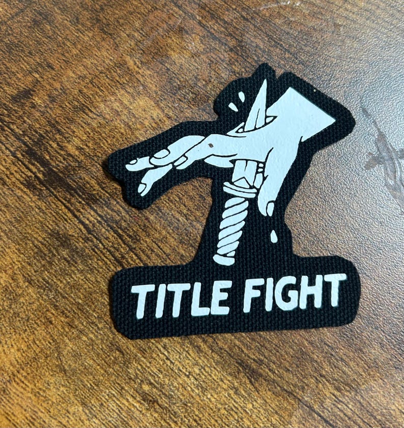 May include: Black and white patch with a hand holding a knife and the text "TITLE FIGHT" underneath.