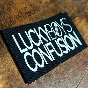 Puede incluir: Parche de tela negro con texto blanco que dice "LUCKYBOYS CONFUSION".