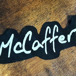 Puede incluir: Texto blanco sobre un parche de tela negro que dice "McCafferty".