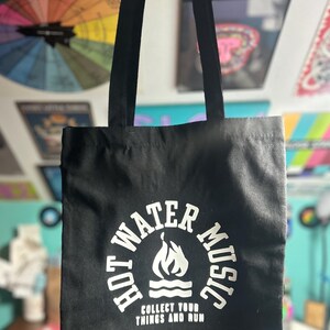 Könnte beinhalten: Schwarze Tragetasche mit dem Text "HOT WATER MUSIC" in einem kreisförmigen Design mit einer Flammen- und Wassergrafik. Der Text "COLLECT YOUR THINGS AND RUN" steht darunter. Die Tasche hat schwarze Griffe.