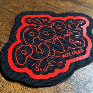Puede incluir: Parche bordado rojo y negro con las palabras "Pop Punks Not Dead" en una fuente de burbujas.