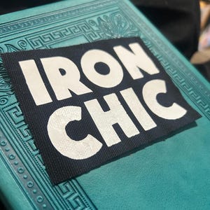 Può includere: Un cerotto in tessuto nero con le parole "IRON CHIC" in lettere bianche in rilievo.