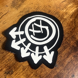 Peut inclure: Patch brodé noir et blanc avec un cercle contenant un smiley avec un 'X' sur les yeux et des flèches pointant vers l'intérieur autour du cercle.