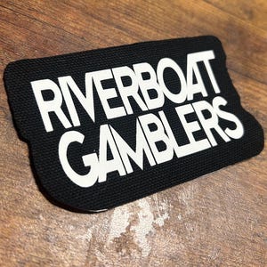 Puede incluir: Un parche de tela negro con texto blanco que dice "Riverboat Gamblers".