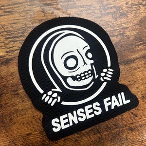 Può includere: Toppa ricamata in bianco e nero con uno scheletro di cartone animato che indossa una tunica con cappuccio e tiene un cerchio. Il testo "SENSES FAIL" è stampato sotto il cerchio.