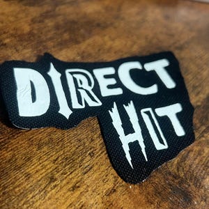 Può includere: Toppa nera e bianca con le parole "DIRECT HIT" in un font stilizzato.