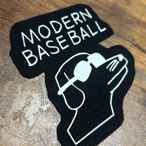 Op de afbeelding: Zwart-witte patch met de tekst "MODERN BASEBALL" en een cartoonhond met een zonnebril.
