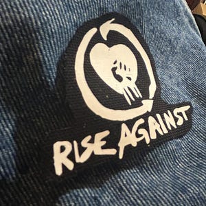 Könnte beinhalten: Ein schwarz-weißes Patch mit einem stilisierten Herz- und Schädelmotiv. Der Text "Rise Against" ist in weißen Buchstaben auf schwarzem Hintergrund geschrieben.