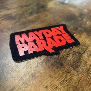 May include: A black patch with the words "MAYDAY PARADE" in bold, red letters. The patch has a rectangular shape with rounded corners. The text is in a stylized font.