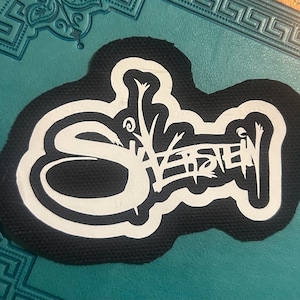 Könnte beinhalten: Schwarz-weiß gestickter Patch mit dem Wort "SVEISTEIN" in einer stilisierten Schrift.