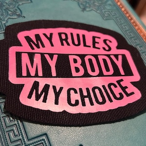May include: A black patch with the text "MY RULES MY BODY MY CHOICE" in pink. The patch has a textured appearance and is set against a teal background with a decorative pattern.