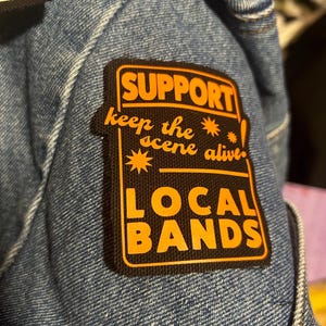 以下が含まれることがあります： 「SUPPORT keep the scene alive! LOCAL BANDS」というテキストが書かれた黒とオレンジの刺繍パッチ。