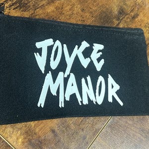 Könnte beinhalten: Schwarze Canvas-Kosmetiktasche mit weißem Schriftzug "JOYCE MANOR".