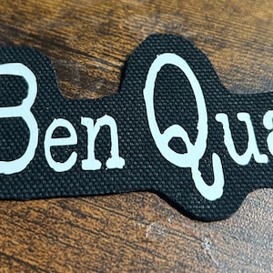 Op de afbeelding: Zwarte stoffen patch met witte tekst "Ben Quad".