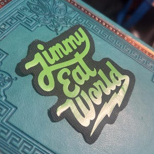 May include: A black and green patch with the text "Jimmy Eat World" in a stylized font. The patch is on a teal surface with a decorative border.