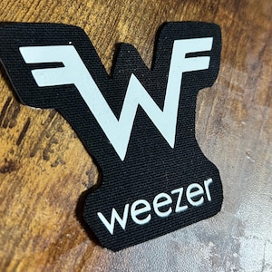 May include: Black patch featuring the band name "Weezer" in white, with a stylized "W" logo. The patch has a rectangular shape with rounded edges. The logo and text are printed in a bold, sans-serif font.