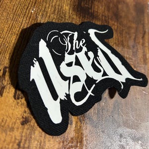 Peut inclure: Patch en tissu noir avec l'inscription "The Used" en blanc dans une police stylisée.